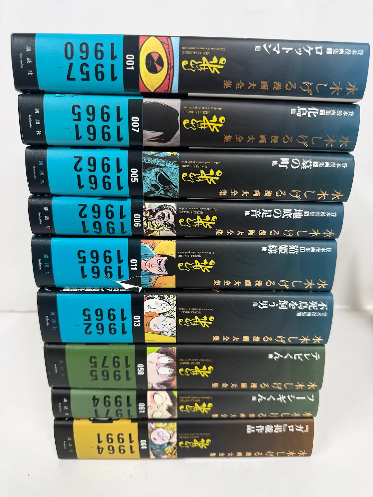 講談社『水木しげる漫画大全集』9冊の背表紙——001巻(1957-1960 ロケットマン他)、005巻(1961-1962 墓の町他)、006巻(1961-1962 地底の足音他)、007巻(1961-1965 化烏他)、011巻(1961-1965 猫姫様他)、013巻(1962-1965 不死鳥を飼う男他)、058巻(1965-1975 テレビくん他)、061巻(1971-1994 フーシギくん他)、064巻(1964-1991 ガロ掲載作品他)。黒背表紙6冊・緑背表紙2冊・黄色背表紙1冊の混在