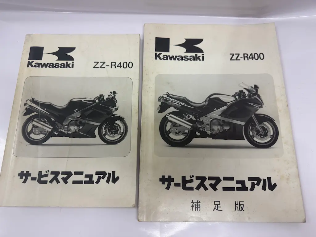 広島市安芸区でお譲りいただいた、カワサキ『ZZ-R400 サービスマニュアル』と『ZZ-R400 サービスマニュアル 補足版』の2冊を並べたところ。どちらも表紙にKawasakiのロゴとZZ-R400の車体の写真が入っており、左が本編、右が補足版。表紙の車体の写真は2冊で異なっている。バイクの雑誌やムック、オートメカニックと一緒にお部屋にあった整備書