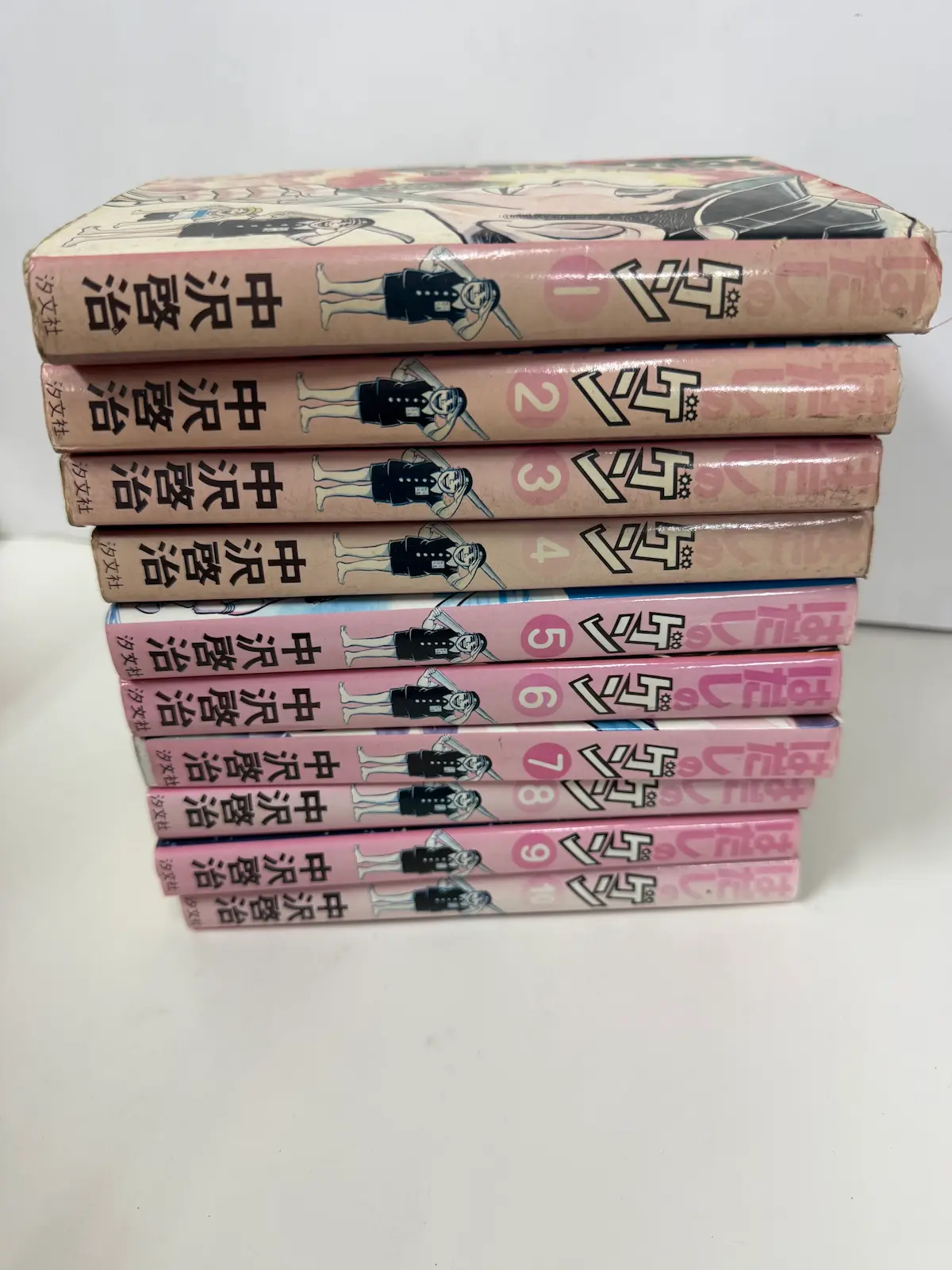 安芸高田市でお譲りいただいた中沢啓治『はだしのゲン』汐文社版 全10巻の背表紙。1巻から4巻は背のピンクが日焼けで抜けてベージュに近い色になり、5巻から10巻はピンクが残っている。各巻の背に学生服姿の少年のイラストと丸囲みの巻数が入り、下部に「汐文社」の社名がある
