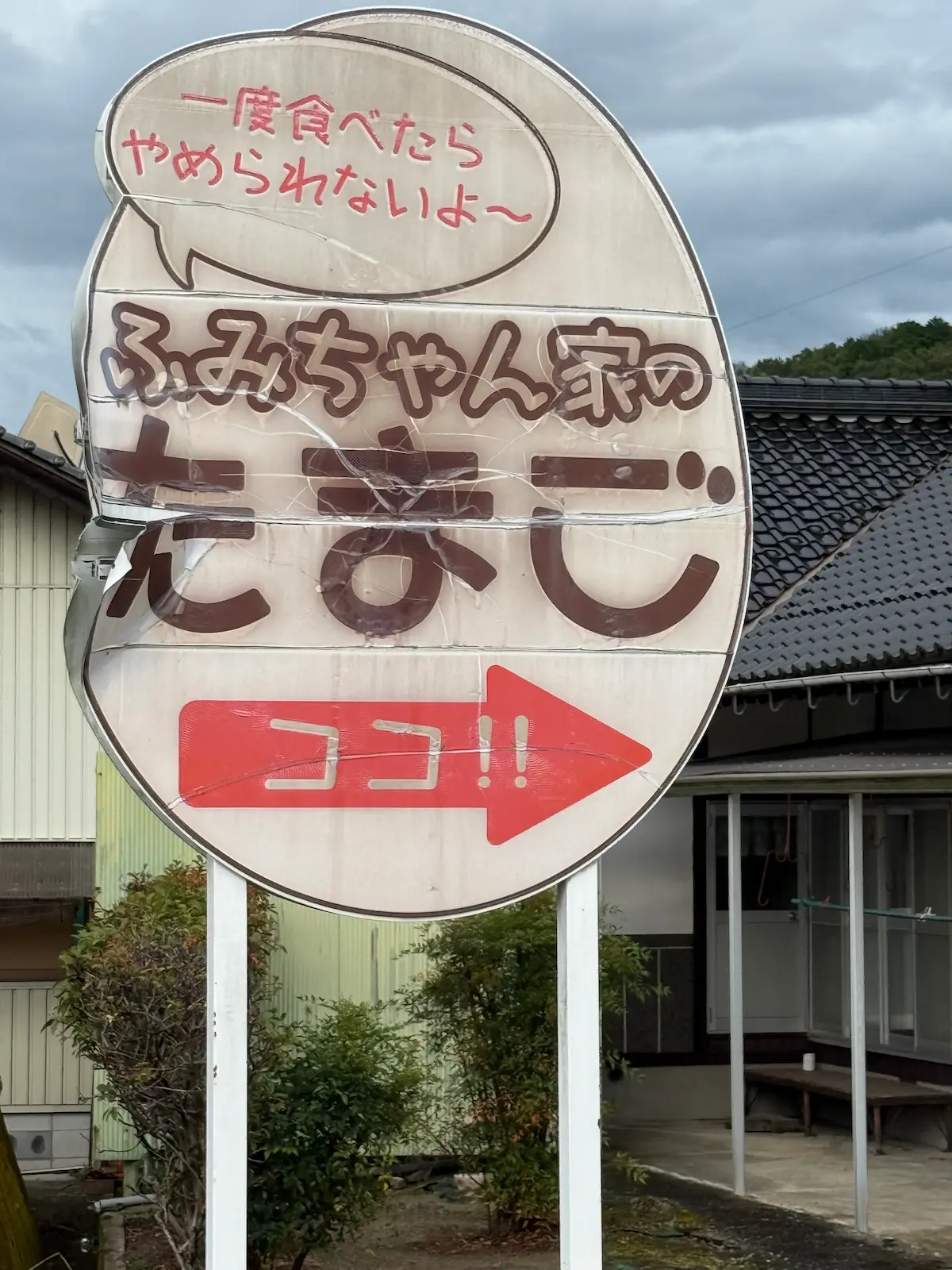 「ふみちゃん家のたまご」と書かれた屋外の看板。上部の吹き出しに「一度食べたら やめられないよ〜」、中央に茶色の丸文字で店名、下部に「ココ!!」と白抜き文字の入った赤い矢印が右を指している。看板にはひび割れと補修跡があり、背景は民家の瓦屋根と木々