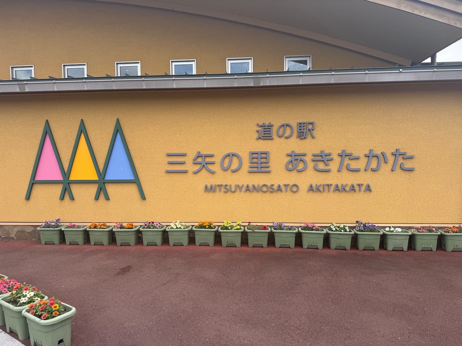 道の駅「三矢の里 あきたかた」の入口看板。「道の駅 三矢の里 あきたかた MICHI-NO-EKI MITSUYANOSATO AKITAKATA」と日本語・ローマ字で大きく表示。横にはAAAを三角形に組み合わせた三本の矢のロゴマーク。背景は新緑の山々と田園風景。
