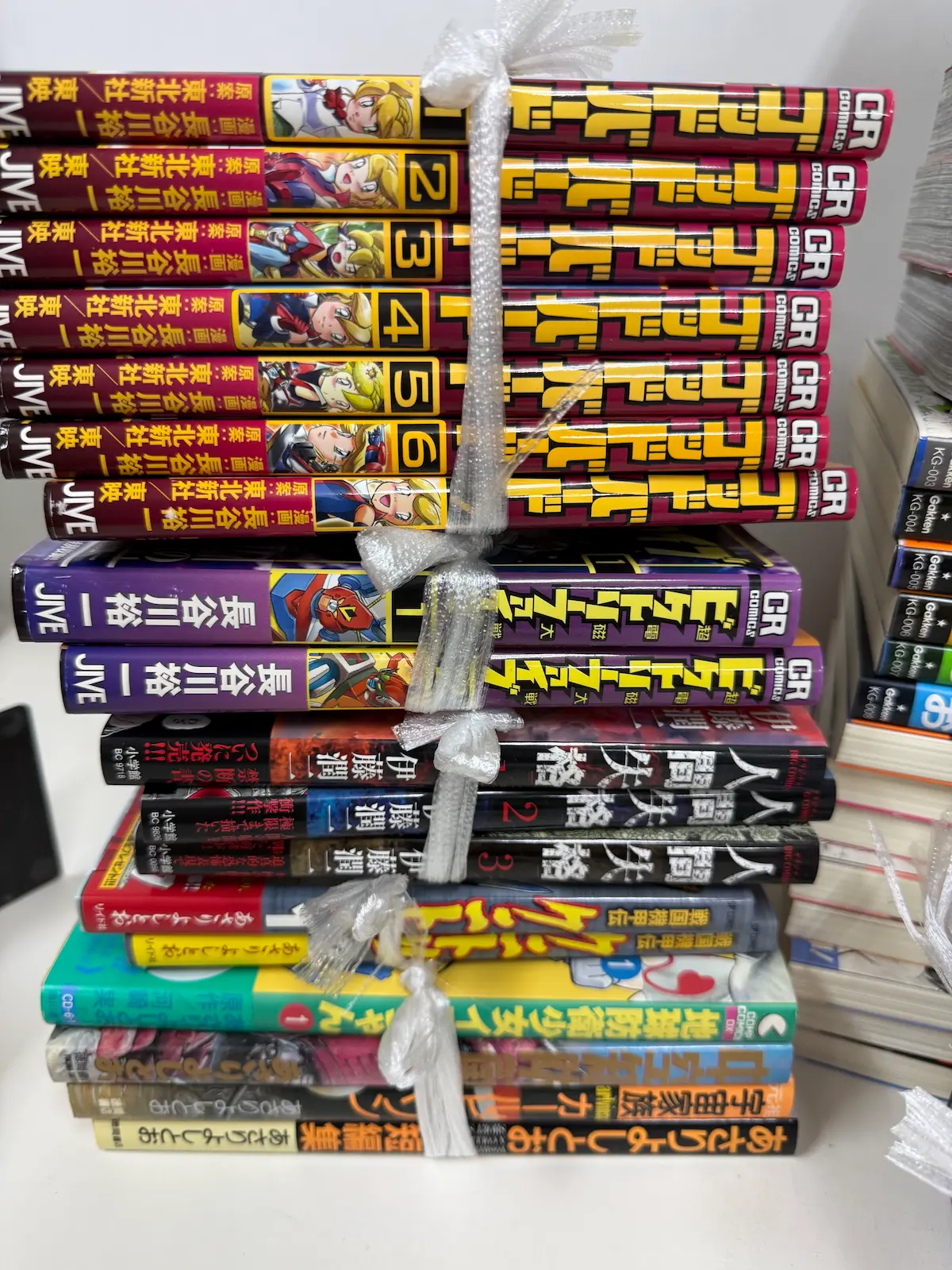 事務所で束にした横積みの背表紙。上から長谷川裕一『ゴッドバード』の深い赤の背が1巻から7巻、JIVE・CR COMICSと原案東映の表記とともに並び、その下に『ビクトリーファイブ』の紫の背2冊、さらに伊藤潤二『人間失格』小学館の黒い背1・2・3巻、一番下にあさりよしとおの単行本が数冊続く