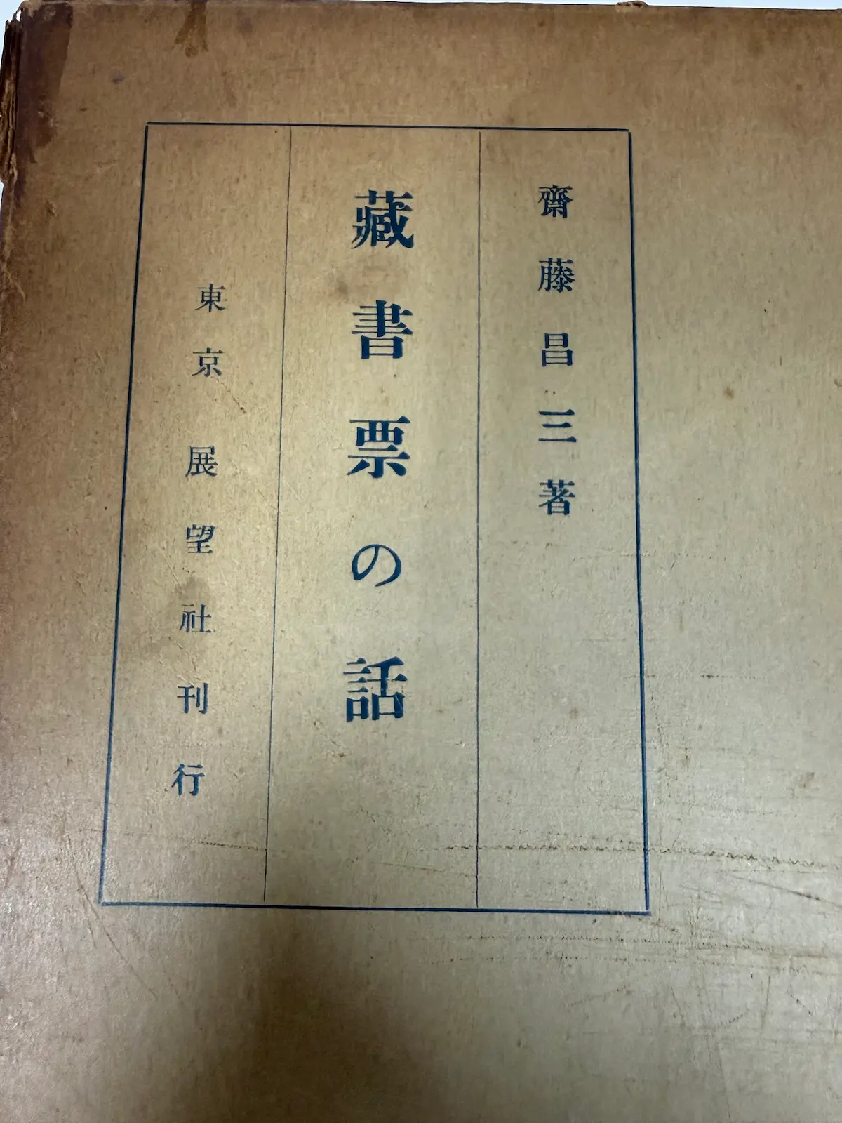 齋藤昌三『藏書票の話』の函の題字部分のアップ。青枠のなかに、右上から下へ縦書きで「齋藤昌三著　藏書票の話　東京　展望社刊行」の題字が入る。函の紙は経年により少しくたびれているが、印字はしっかり残っている
