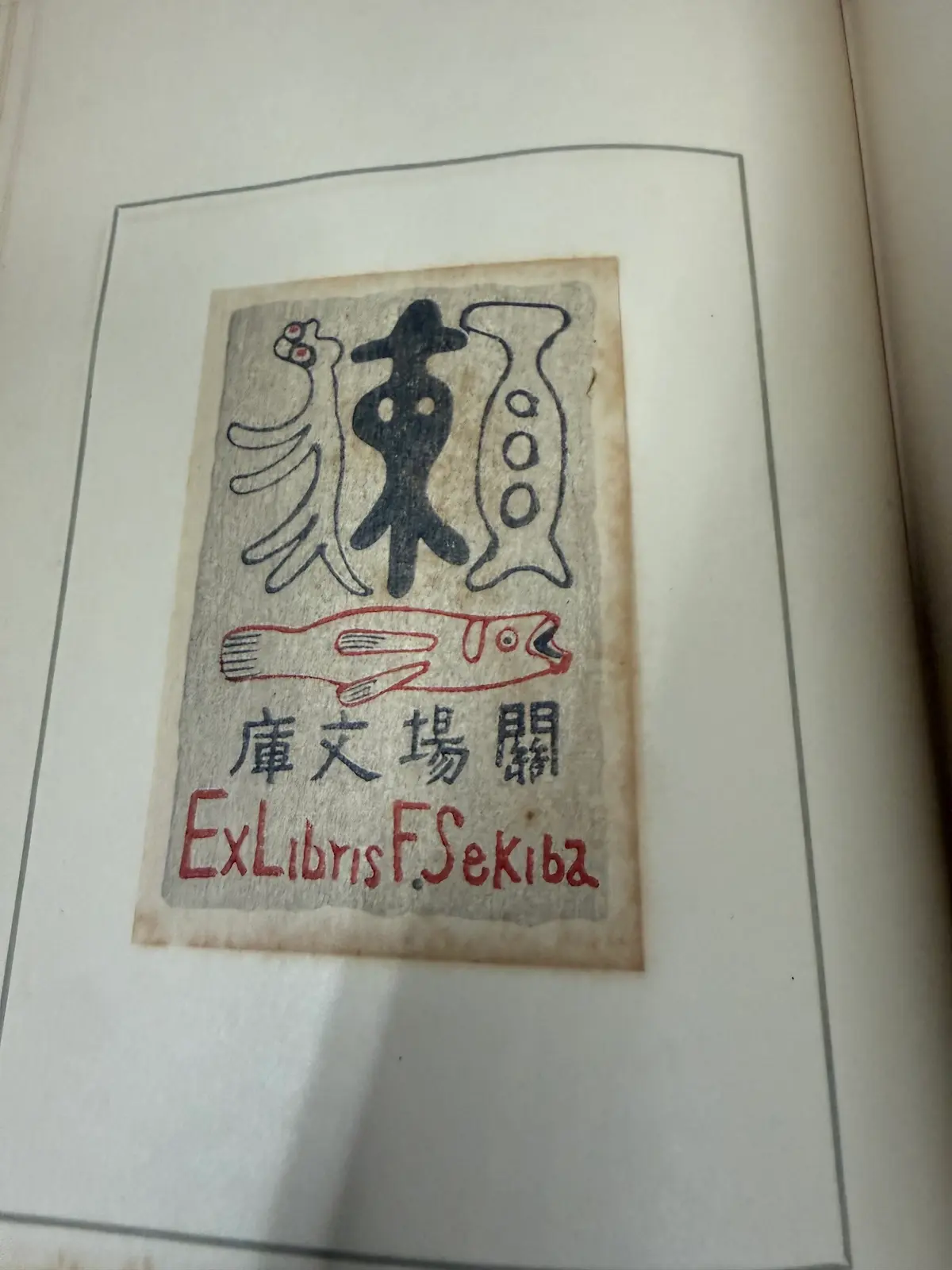 『藏書票の話』のページに実物で貼り込まれた蔵書票の一枚。朱色の魚と、その下に甲骨文字風の意匠、上部に「關場文庫」、下部に「F.Sekiba」の英字署名。木版の刷りで、朱と黒の二色刷り