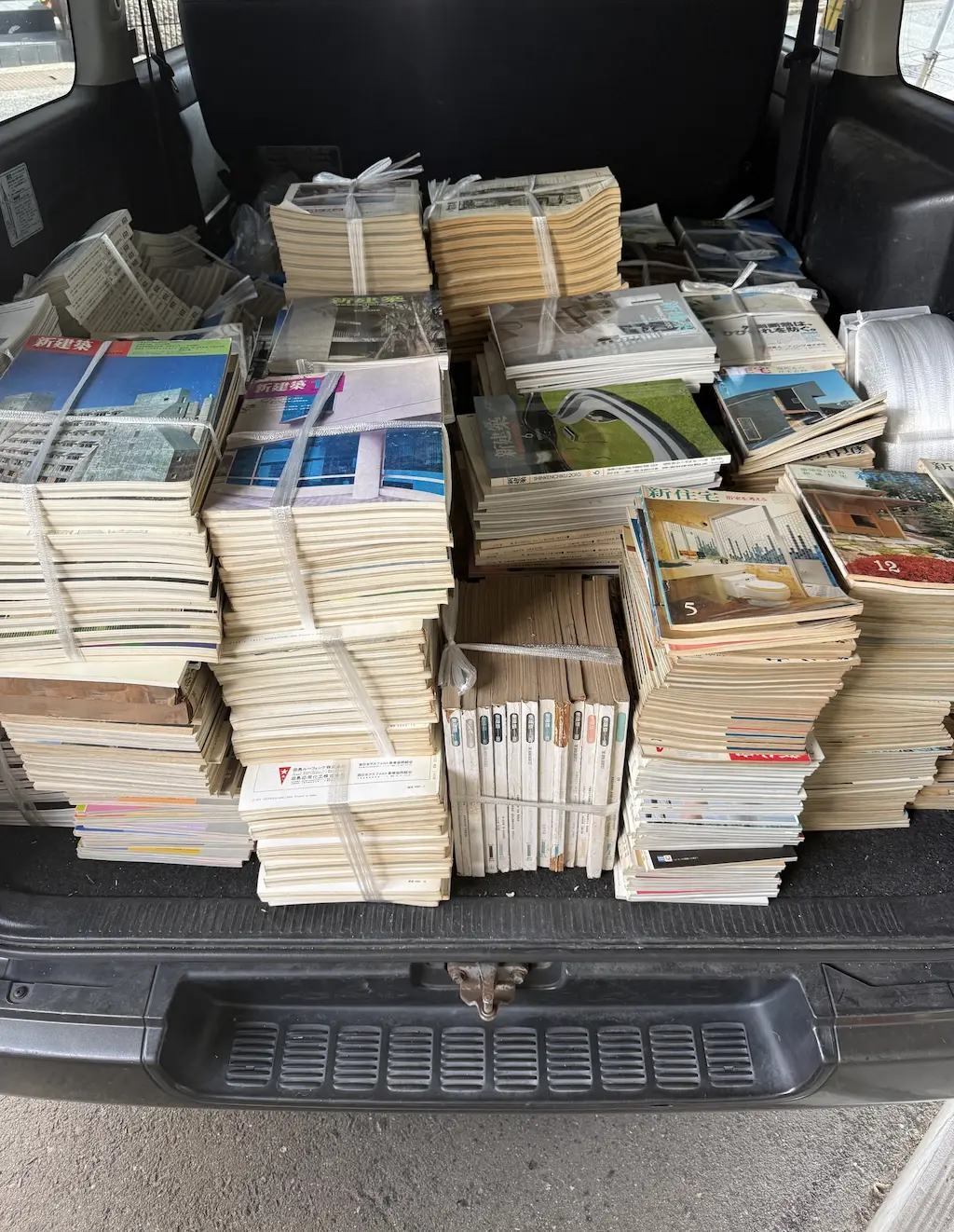 廿日市市でお譲りいただいた建築雑誌を、当店のハイエースの荷室に積み込んだところ。紐で縛った束がいくつも積まれ、手前左の束の上には新建築社『新建築』の表紙、右側には新住宅社『新住宅』の12月号（表紙に「和風住宅」の文字）が見える。中央手前には、背の色が古い『新建築』を立てて縛った束がある。2回目のお引き取りの分