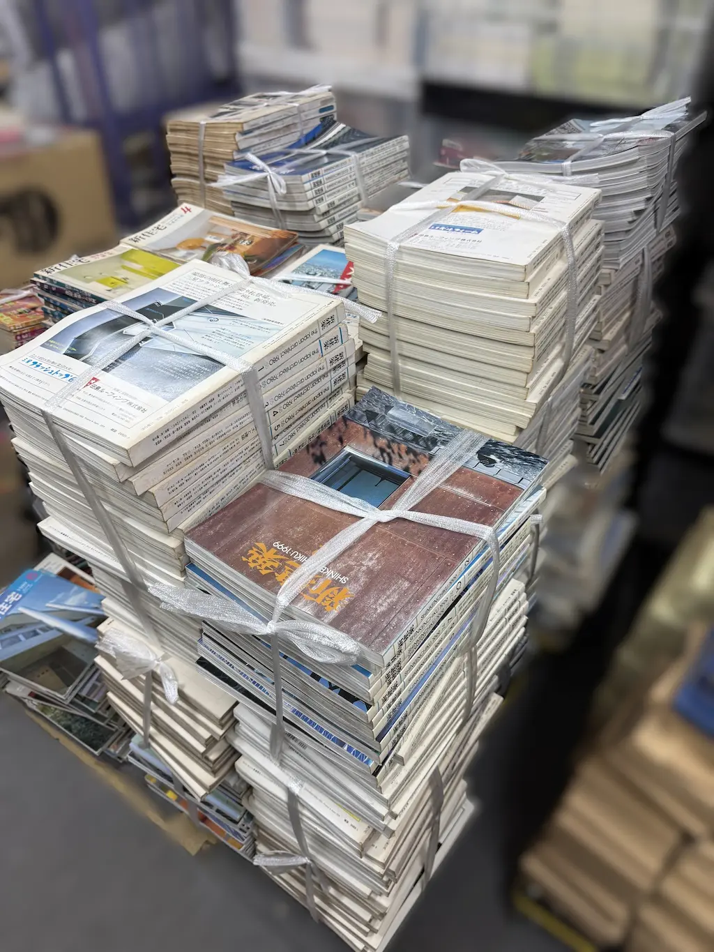 廿日市市でお譲りいただいた新建築社『新建築』の束を、当店事務所で積み上げたところ。手前の束は背に「SHINKENCHIKU:1999」とある1999年の号、左の束は背に「the japan architect:1980」と1から6の番号が並ぶ1980年1月号から6月号。左奥には新住宅社『新住宅』4月号の表紙も見える。どの束も紐で縛ってある