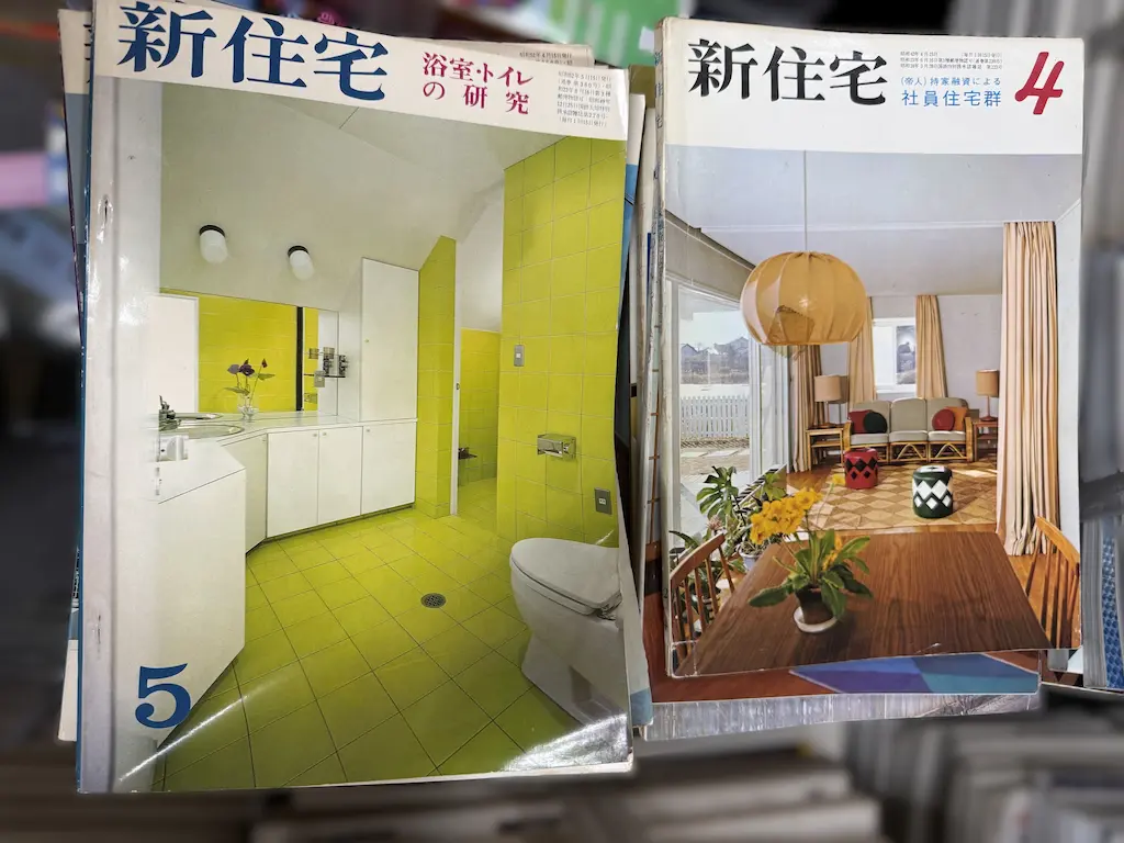 廿日市市でお譲りいただいた新住宅社『新住宅』の2冊を並べたところ。左は昭和52年（1977年）5月号、特集「浴室・トイレの研究」で、黄緑色のタイルの浴室とトイレの写真が表紙。右は昭和42年（1967年）4月号、「（帝人）持家融資による社員住宅群」で、丸いペンダントの照明、籐のソファ、赤と緑のスツールが並ぶ居間の写真が表紙