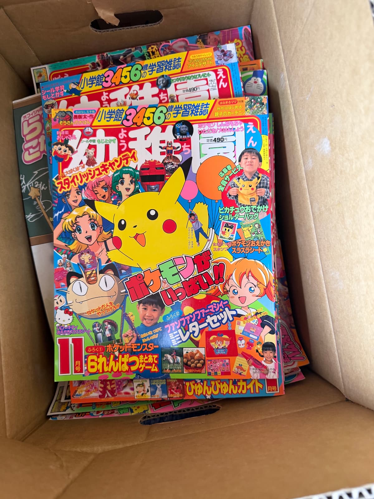 段ボールに入った状態の小学館「幼稚園」90年代後半号。表紙にピカチュウ・ニャース・トゲピーといったポケモン初代から金銀過渡期のキャラクター、ファンファンファーマシィー、ハローキティ、ドラえもんなどが配される。「ポケットモンスター 6れんぱつまとあてゲーム」「ピカチュウおでかけショルダーバッグ」などの付録告知。定価490円。