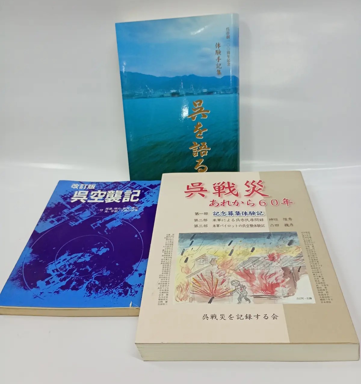アッシュ書店の郷土史の棚に並ぶ呉空襲関連の証言集三冊。左手前『改訂版 呉空襲記』（濃紺表紙、空襲被害の航空写真に同心円マーキング、付・尾道、福山、岩国、徳山、光、宇部、下関の被爆）、右手前『呉戦災 あれから60年』（呉戦災を記録する会編、焼夷弾を浴びる家屋を描いた表紙水彩画、古江町・佐藤氏画）、後方『呉を語る 体験手記集』（呉市制100周年記念、呉湾と灰ヶ峰・造船クレーンの写真に金文字）。