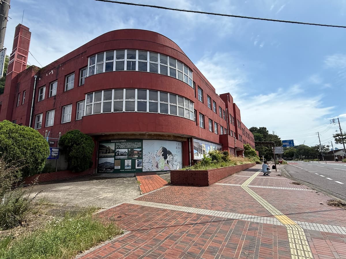 解体直前の青山クラブを店主が現地撮影した全景写真。2026年6月、赤茶色のレンガ調外壁、特徴的なR形状（半円形カーブ）のファサードが高い青空を背に美しく聳えている。三階建て、横長連続窓、陸屋根、左端に煙突状の点迫部。手前にレンガ調の歩道と植込、道路と【入船山】・【呉駅】表示の道路標識が見える。
