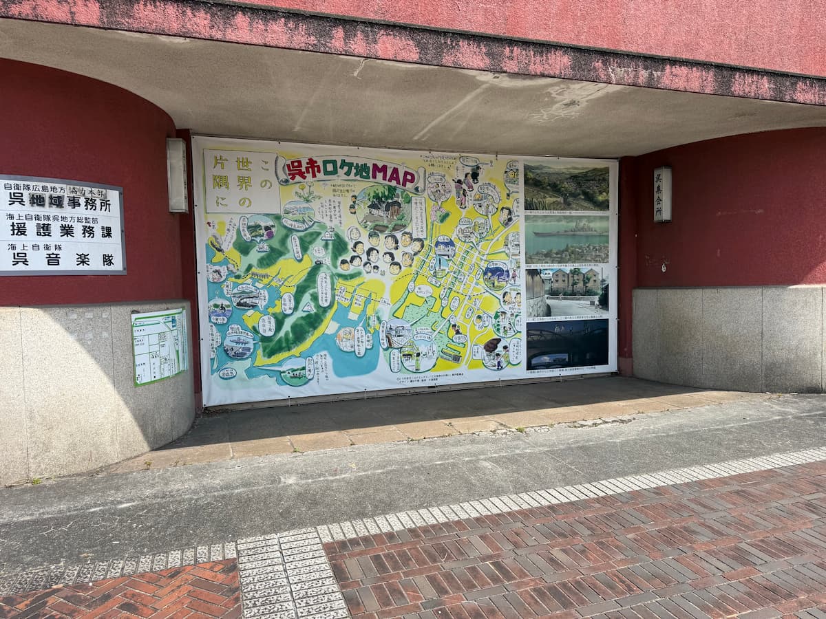 青山クラブ正面アーチに掲示された『この世界の片隅に』呉市ロケ地MAPの現地写真。カラフルなイラストMAPにコマの表情、呈子茶色の魚介、外・休山・長代・などの地名・イラスト・右側に昭和期呉のロケ地写真4枚。左側に「自衛隊広島地方協力本部 呉地域事務所」「海上自衛隊呉地方総監部 援護業務課」「海上自衛隊 呉音楽隊」の3枚の黒字プレート、右側に「呉集会所」のプレートが掛けられている。赤茶色のレンガ調壁面のアーチの内側に設置されている。
