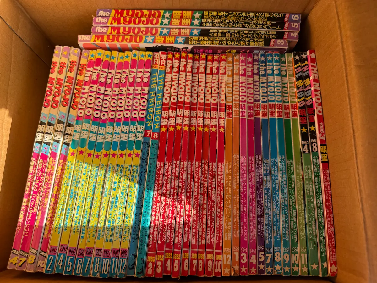 呉市でお譲りいただいた1980年から1984年の『明星』『平凡』50冊を、段ボール箱に背を上にして並べた状態。ピンク、赤、オレンジ、緑と年ごとに背の色が変わり、下端に発行年と月号が印刷されている。奥に数冊が平積みになっている