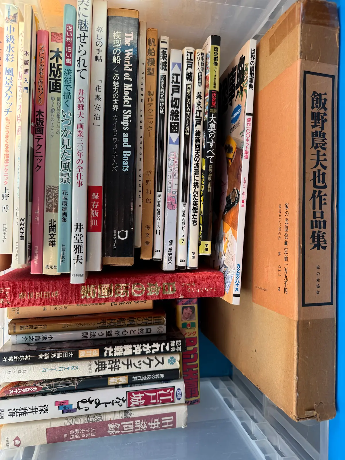 呉市でお譲りいただいた本を青いオリコン（折り畳みコンテナー）に立てて並べた状態。左に木版画・水彩のテクニック本、中央に帆船模型と江戸城・幕末の本、右に函入りの大型本『飯野農夫也作品集』（家の光協会）が立っている。手前には横積みの本が重なっている