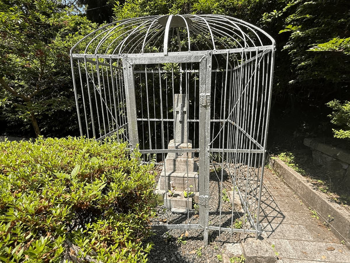 呉海軍墓地（長迫公園）に建つ、銀色の鉄柵ドームに守られた英国水兵GEORGE TIBBINSの墓。十字架の墓石を金属製の檻のような鉄柵が完全に覆っており、奥には深い緑の森が広がっている。