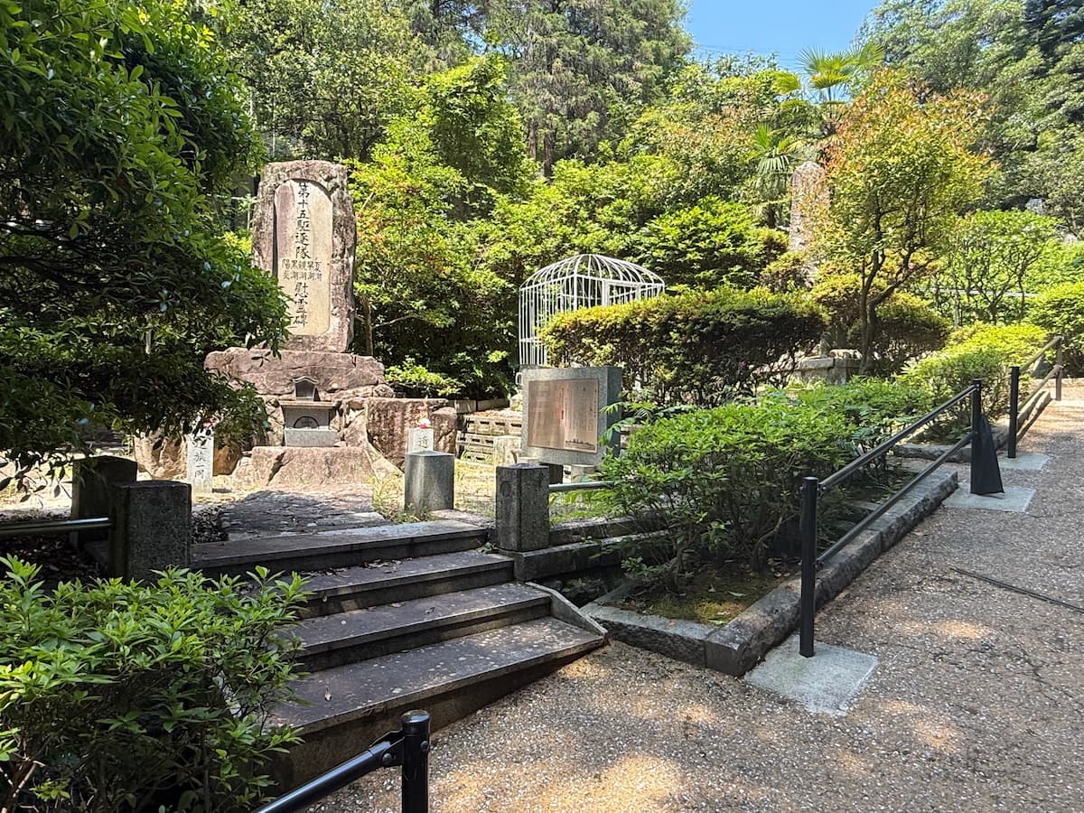 長迫公園内、TIBBINSの墓と背後の慰霊碑群の位置関係。中央奥にシルバーのドーム状鉄柵に囲まれた英国水兵の墓が見え、その左隣の階段の上には大きな石碑（第十五駆逐隊慰霊碑）がそびえる。