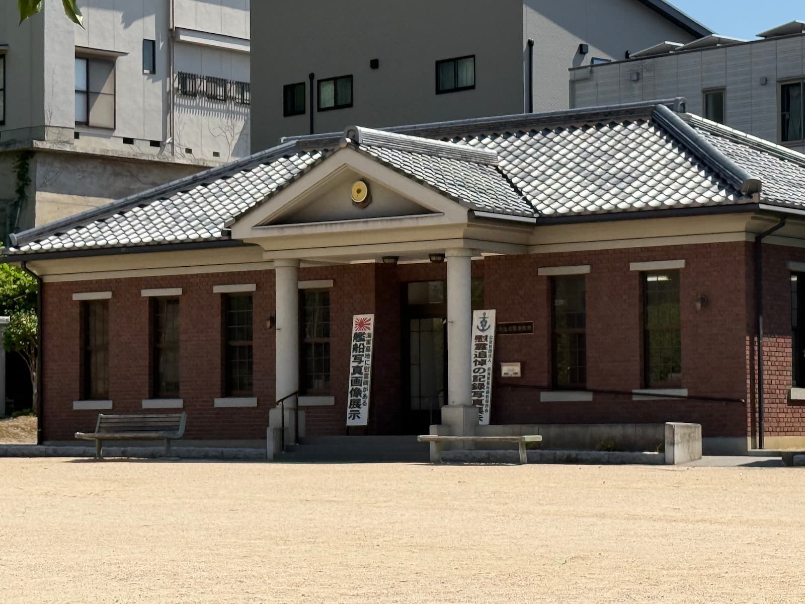 長迫公園内に建つ公益財団法人 呉海軍墓地顕彰保存会の事務局。赤レンガと和瓦の屋根を持つ和洋折衷の小さな建築で、入口には「艦船写真画像展示」「殉難追悼の記録写真展」などの幟が立てられている。