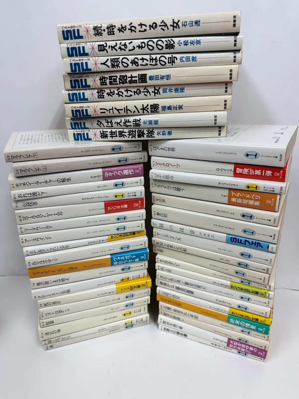 お譲りいただいたSF蔵書の全体。上段に鶴書房SFベストセラーズのソフトカバー8冊が横積みされ、その下に文庫本が2列に積み上がっている。文庫の背表紙にはサンリオSF文庫特有の背番号とマークが見える