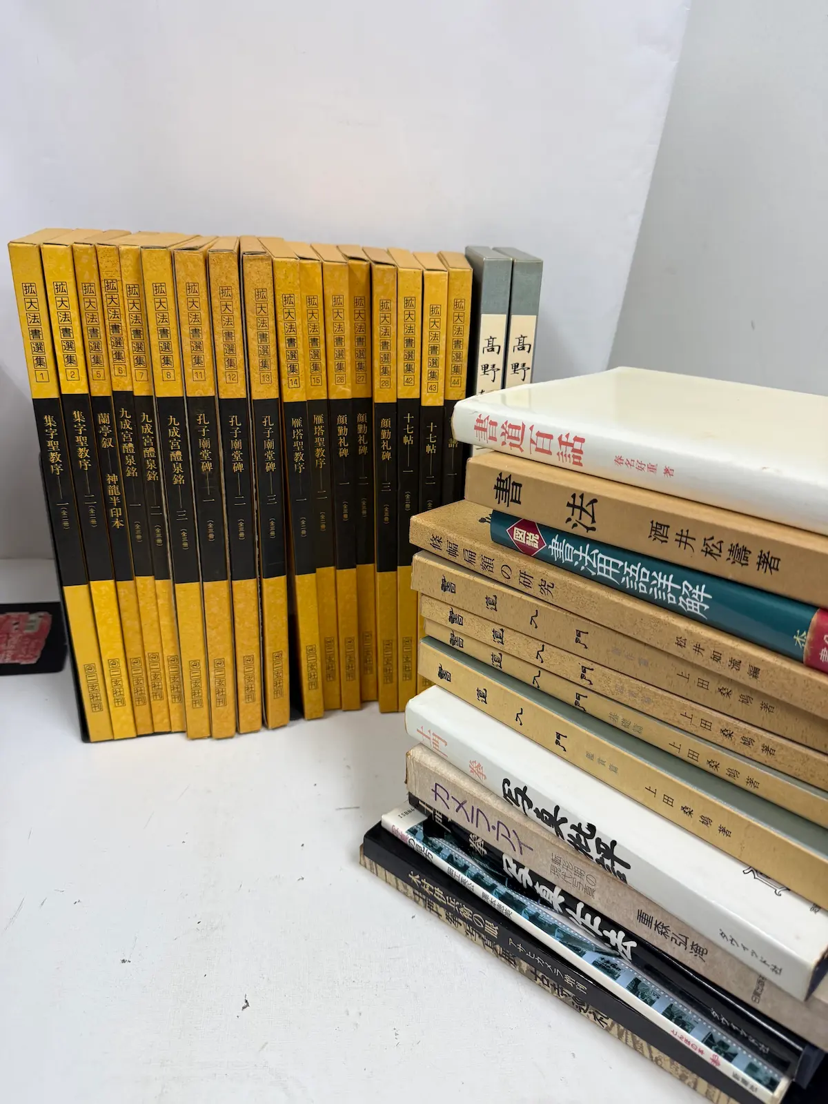 呉市内でお譲りいただいた書道関係と写真関係の本、計33冊を並べたところ。奥に二玄社『拡大法書選集』の背表紙がずらりと立ち並び、右手にグレーの函に入った『拡大かな選集』高野切、手前に写真関係の単行本と書道の入門書が積まれている