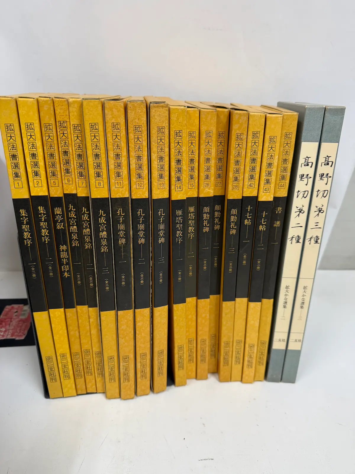 呉市内でお譲りいただいた二玄社『拡大法書選集』17冊の背表紙が左から順に並んだところ。背の上部に巻番号を囲む四角い枠があり、1・2の集字聖教序、5の蘭亭叙 神龍半印本、6・7・8の九成宮醴泉銘、11・12・13の孔子廟堂碑、14・15の雁塔聖教序、26・27・28の顔勤礼碑、42・43の十七帖、44の書譜が続き、右端にグレーの函に入った『拡大かな選集』高野切 第二種・第三種が立っている