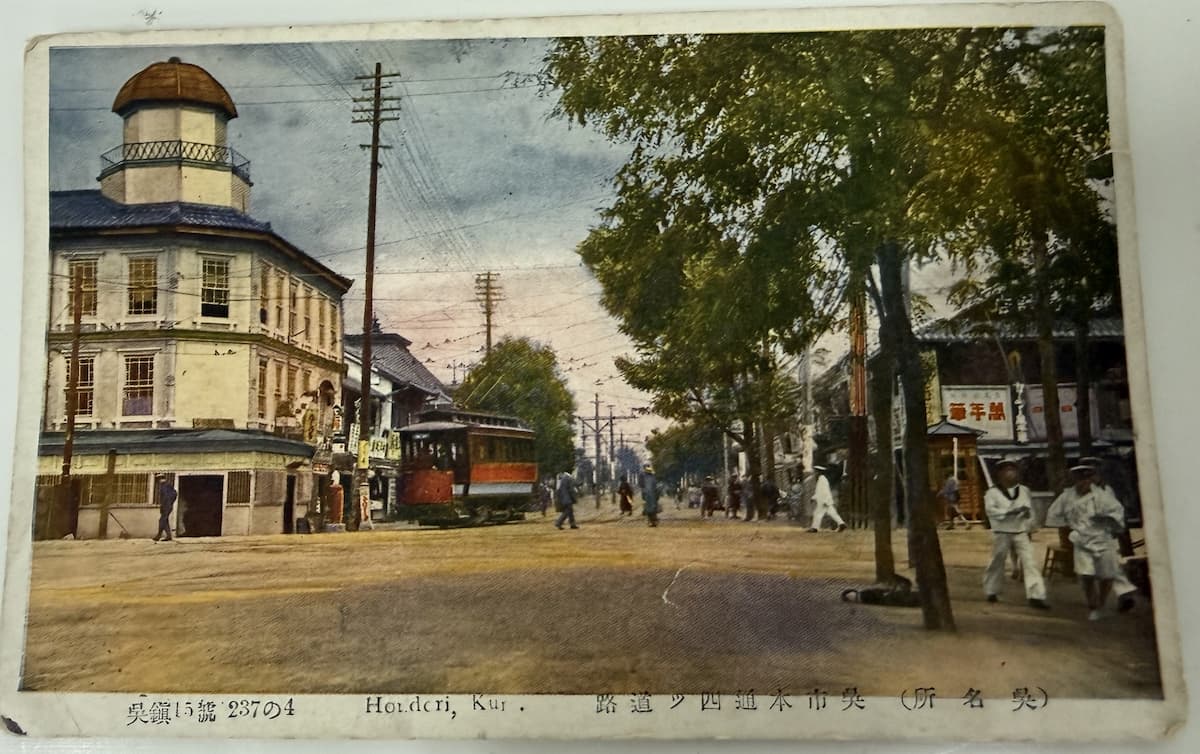 戦前の呉市本通リ四ツ道路を写した手彩色絵はがき。左側に三階建てのドーム屋根の時計屋洋館、中央に呉市電（赤とクリーム色の路面電車）、右側に街路樹と海軍水兵（白い制服）2名を含む通行人が写る。下部キャプション「呉鎭15號 237の4／Hondori, Kure.／呉市本通四ツ道路（呉名所）」。