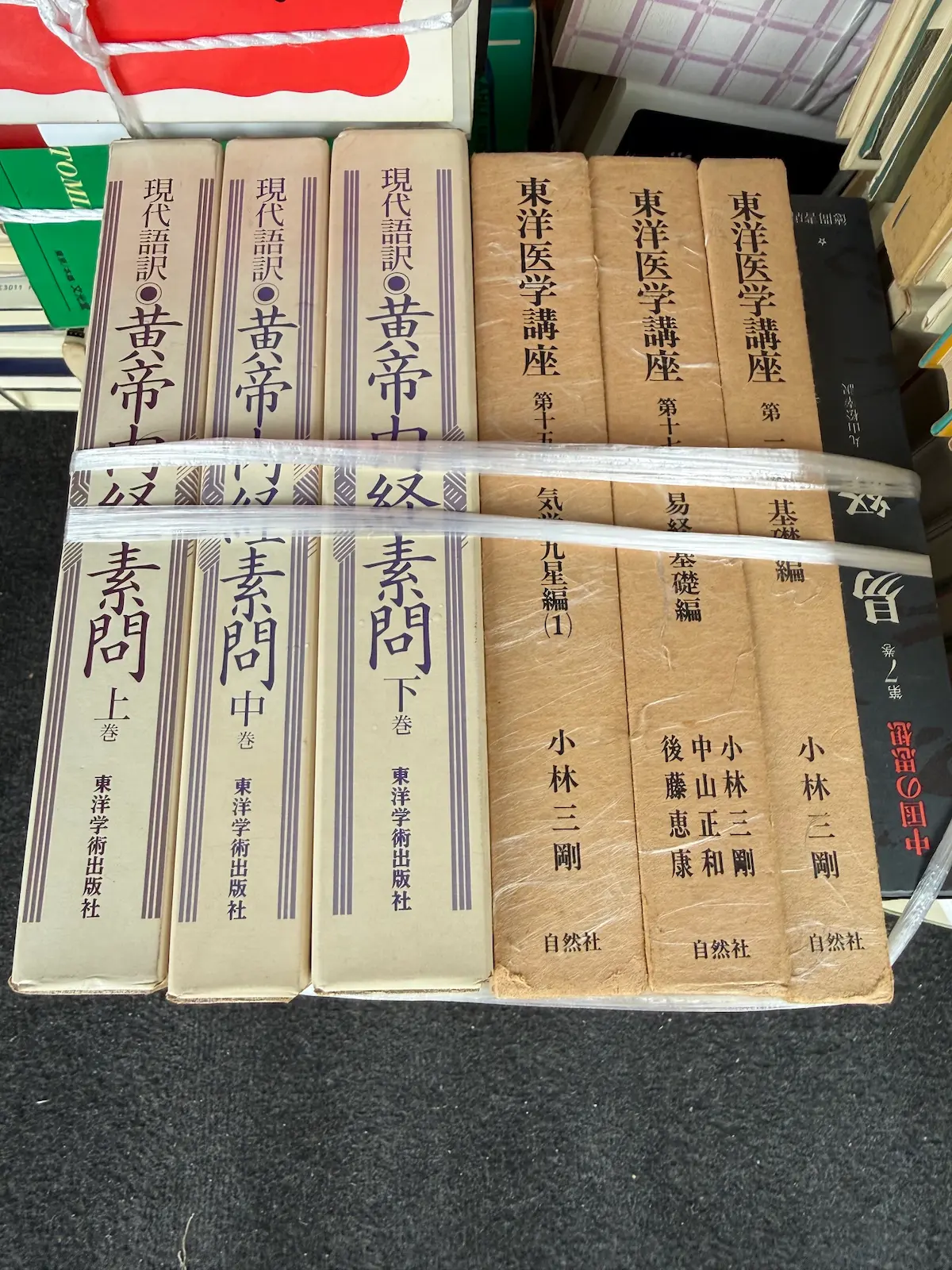 函入りの東洋医学書7冊を平置きで並べた状態。左に東洋学術出版社『現代語訳 黄帝内経 素問』の上巻・中巻・下巻の3冊が揃って並び、右に自然社『東洋医学講座』小林三剛の第十五巻 気学九星編（1）、第十四巻 易経 基礎編、第一巻 基礎編の3冊が並ぶ。東洋医学講座の函にはビニールカバーが残っている。右端に『中国の思想 第7巻 易』