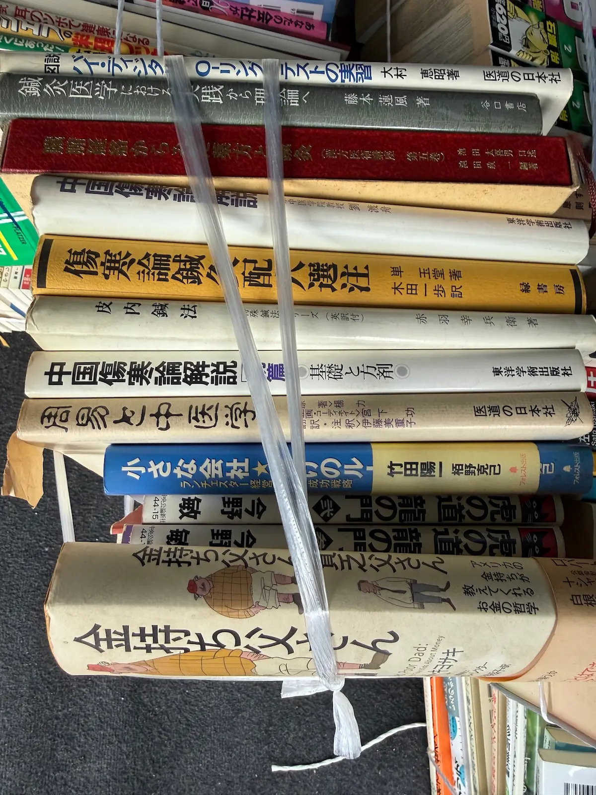 鍼灸・中医学の専門書を横積みにした束の背表紙。上から大村恵昭『バイ・デジタル O-リングテストの実践』医道の日本社、藤本蓮風『鍼灸医学における実践から理論へ』谷口書店、尾台榕堂『類聚方広義』漢方医書集成、『中国傷寒論解説』前篇・後篇 東洋学術出版社、単玉堂『傷寒論鍼灸配穴選注』緑書房、赤羽幸兵衛『皮内鍼法』が並び、下部にビジネス書が混じる。半透明のPPバンド2本で十文字に縛られている