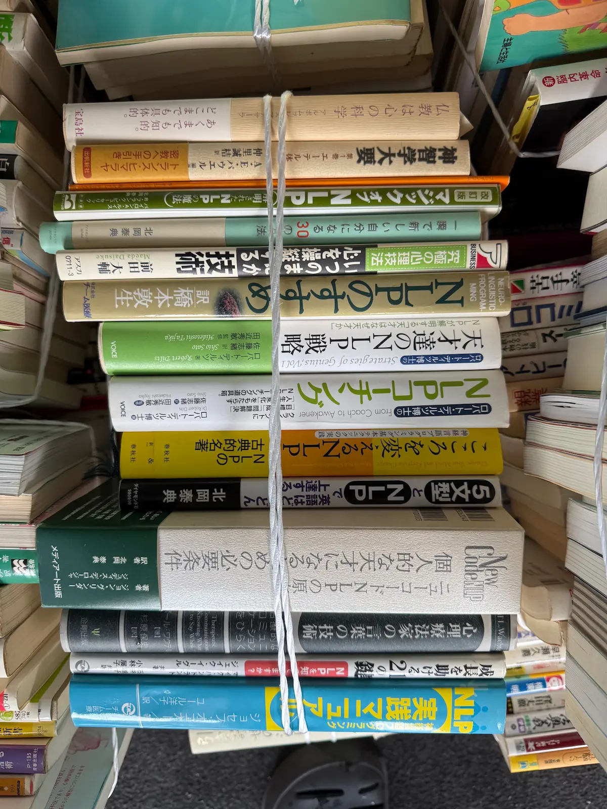 NLPと心理療法の単行本を縦に積んだ束の背表紙。ロバート・ディルツ『天才たちのNLP戦略』『NLPコーチング』VOICE、ジョン・グリンダーのニューコードNLP関連（春秋社）、北岡泰典の著書、ジョセフ・オコナー『NLP実践マニュアル』チーム医療、アーサー・E・パウエル『神智学大要 第一巻 エーテル体』などが並び、白いビニール紐1本で縛られている