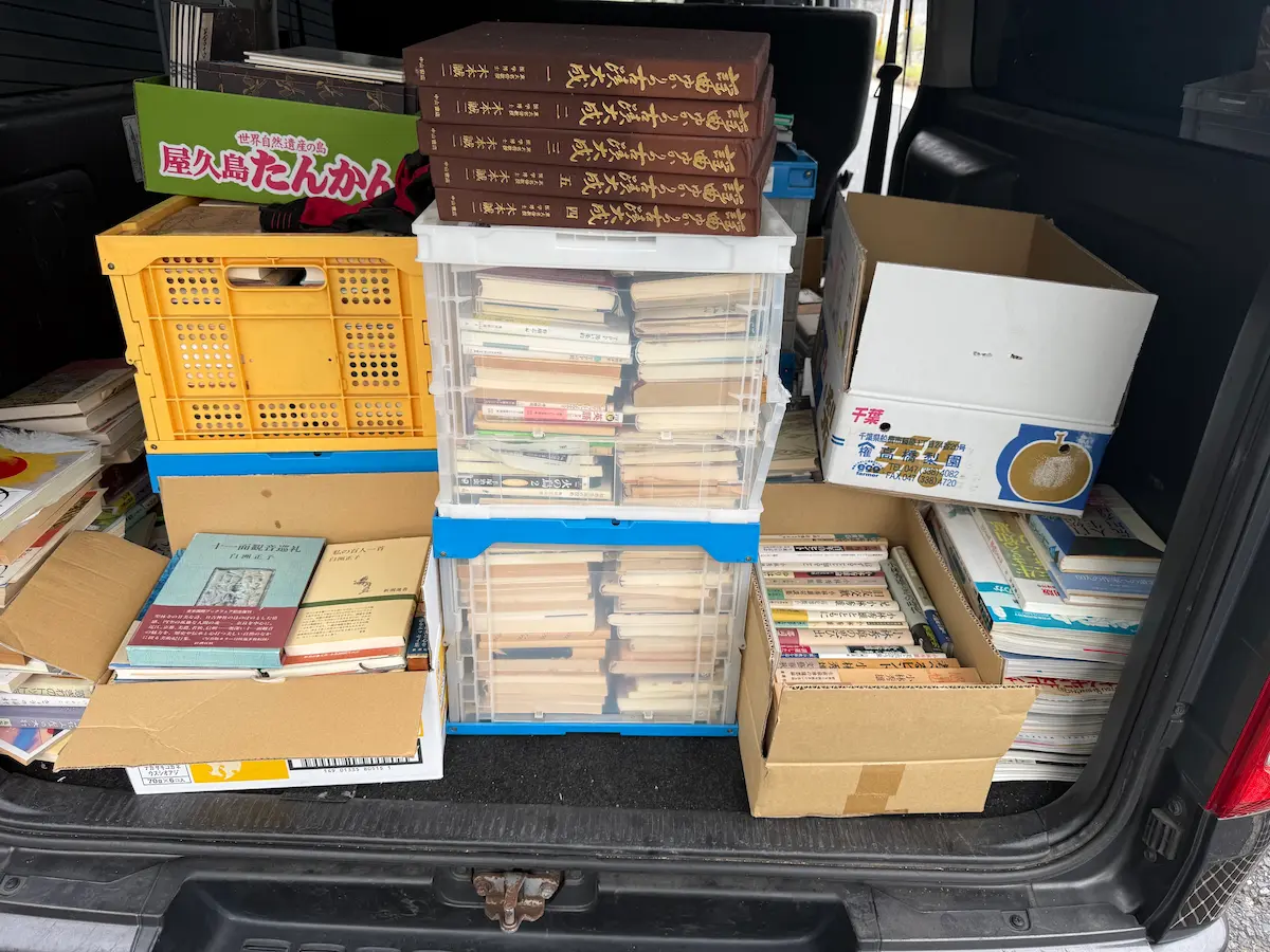 広島市西区の蔵書整理でお預かりした書籍を車の荷室に積み終えた様子。黄色いオリコン、透明の折りたたみコンテナ、段ボール箱が積み重なり、上には茶色い函入りの5冊セットが平積みで置かれている。オリコン8箱分