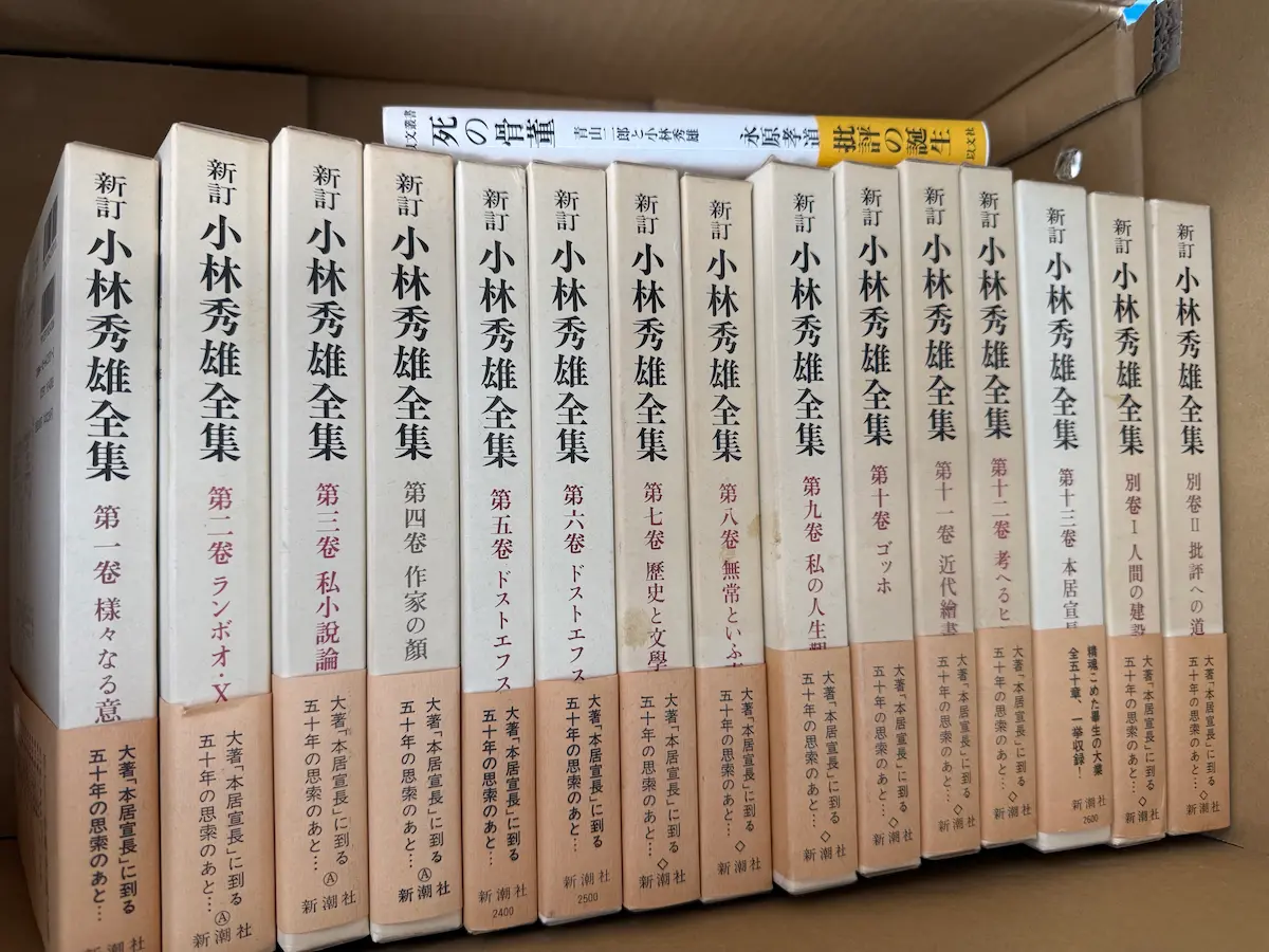 新潮社『新訂 小林秀雄全集』全15冊を段ボール箱に立てて並べた状態。第一巻『様々なる意匠』から第十三巻『本居宣長』、別巻Ⅰ『人間の建設』、別巻Ⅱ『批評への道』までの背表紙が並び、帯には「大著『本居宣長』に到る五十年の思索のあと」と記されている