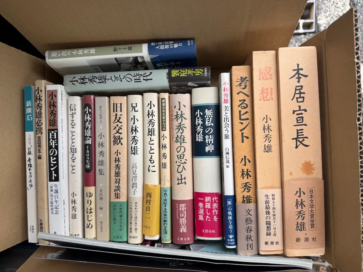 小林秀雄に関する書籍16点を段ボール箱に立てて並べた状態。吉田熈生編『小林秀雄必携』（學燈社）、『小林秀雄 百年のヒント』（新潮 四月臨時増刊）、高見澤潤子『兄 小林秀雄』、郡司勝義『小林秀雄の思ひ出』、『無私の精神』、『考えるヒント』、『感想』、函入りの『本居宣長』などの背表紙が並んでいる