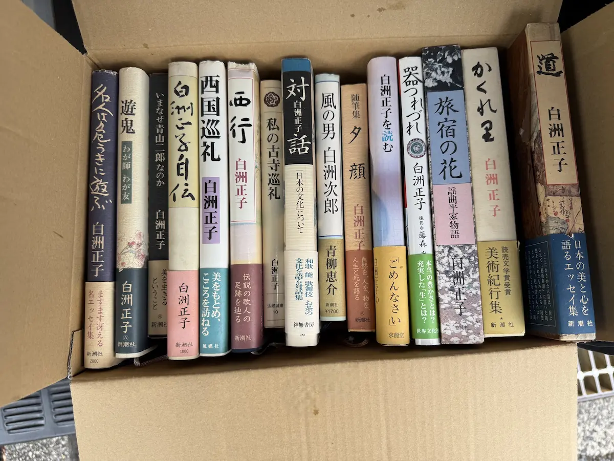 白洲正子の著書15点を段ボール箱に立てて並べた状態。『名人は危うきに遊ぶ』『遊鬼 わが師わが友』『いまなぜ青山二郎なのか』『白洲正子自伝』『西国巡礼』『西行』『私の古寺巡礼』（法藏館）『対話 白洲正子 日本の文化について』（神無書房）、青柳恵介『風の男 白洲次郎』、『随筆集 夕顔』『器つれづれ』『旅宿の花 謡曲平家物語』『かくれ里』、函入りの『道』などの背表紙が並んでいる